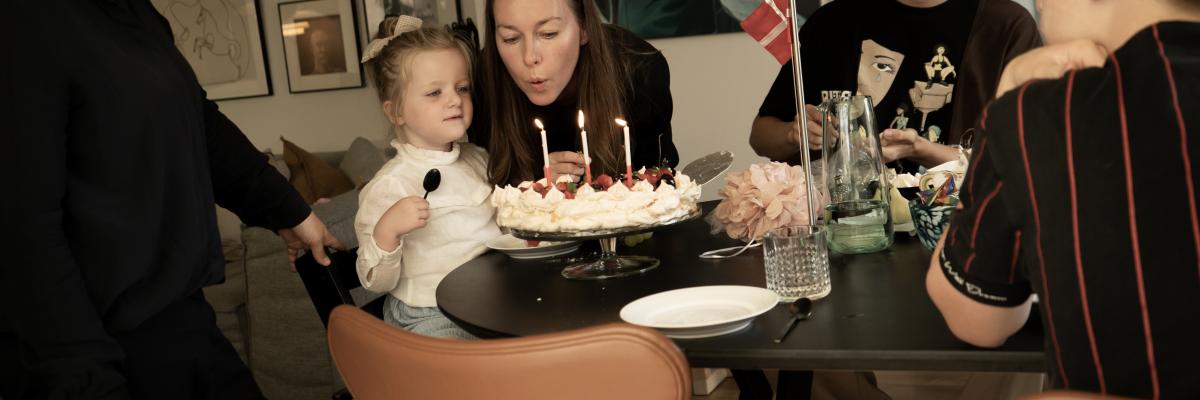 Familie sidder rundt om bordet. Pige puster lys ud på en fødselsdagslagkage.